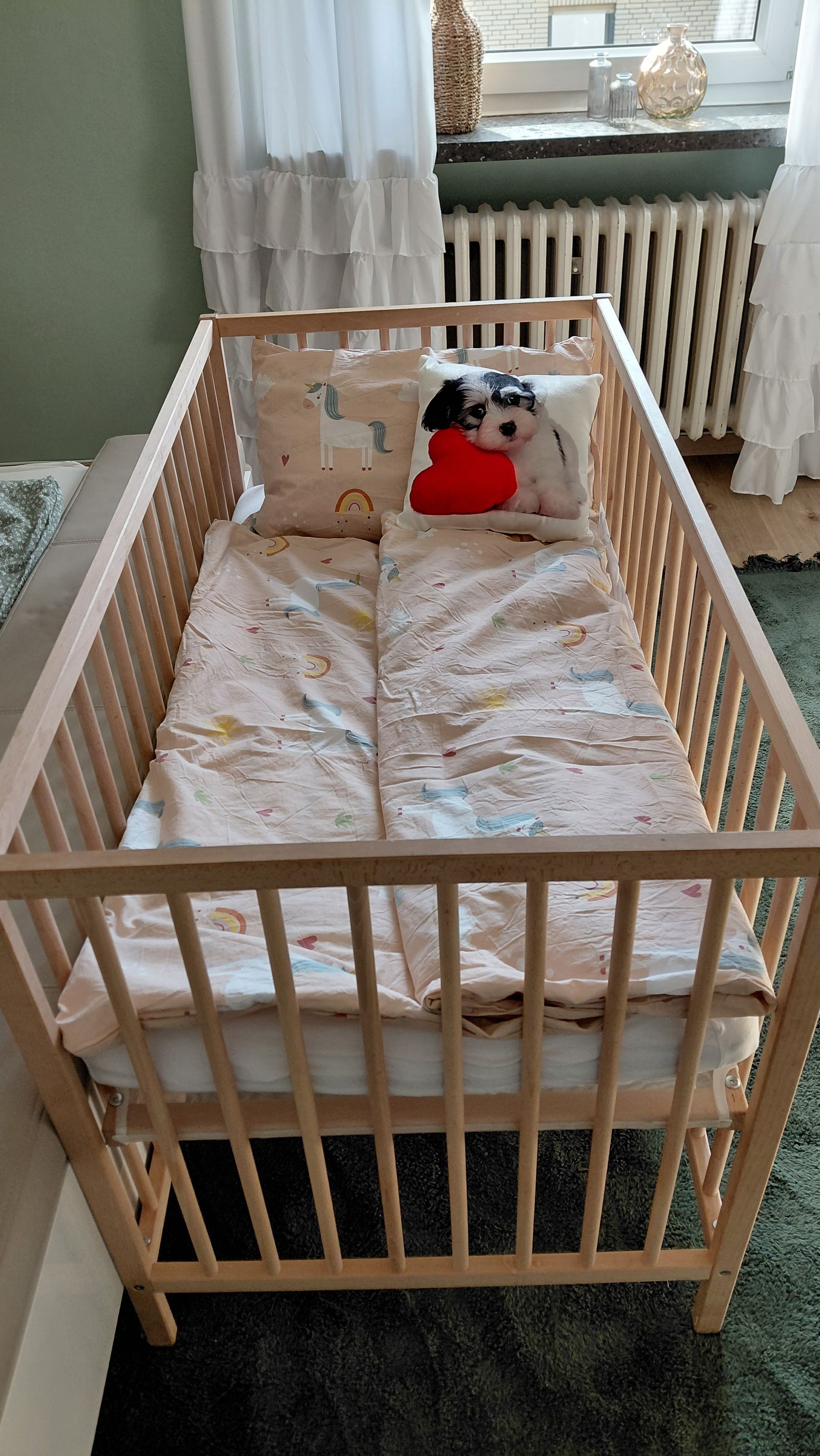 A wooden crib with a patterned mattress, a red heart pillow, and a unicorn pillow.