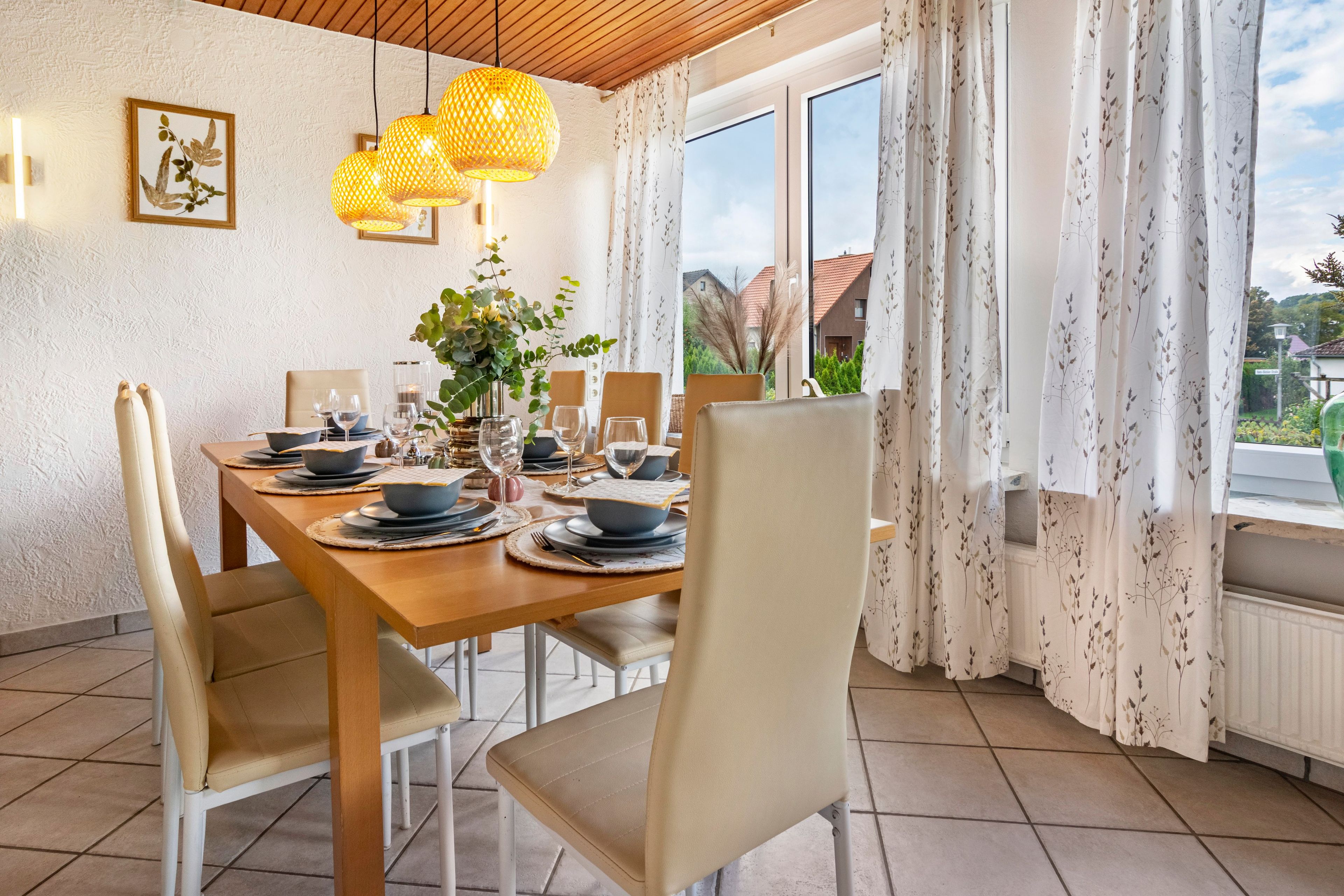 A dining room with a wooden table set for four, surrounded by chairs, with a view of greenery outside.