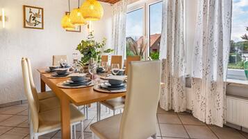 A dining room with a wooden table set for four, surrounded by chairs, with a view of greenery outside.