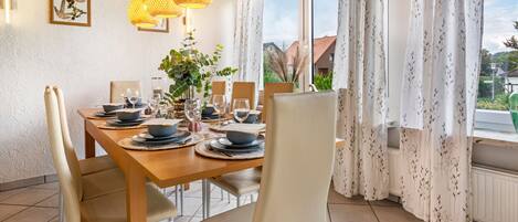 Une salle à manger avec une table en bois dressée pour quatre personnes, entourée de chaises, et offrant une vue sur la verdure à l’extérieur.