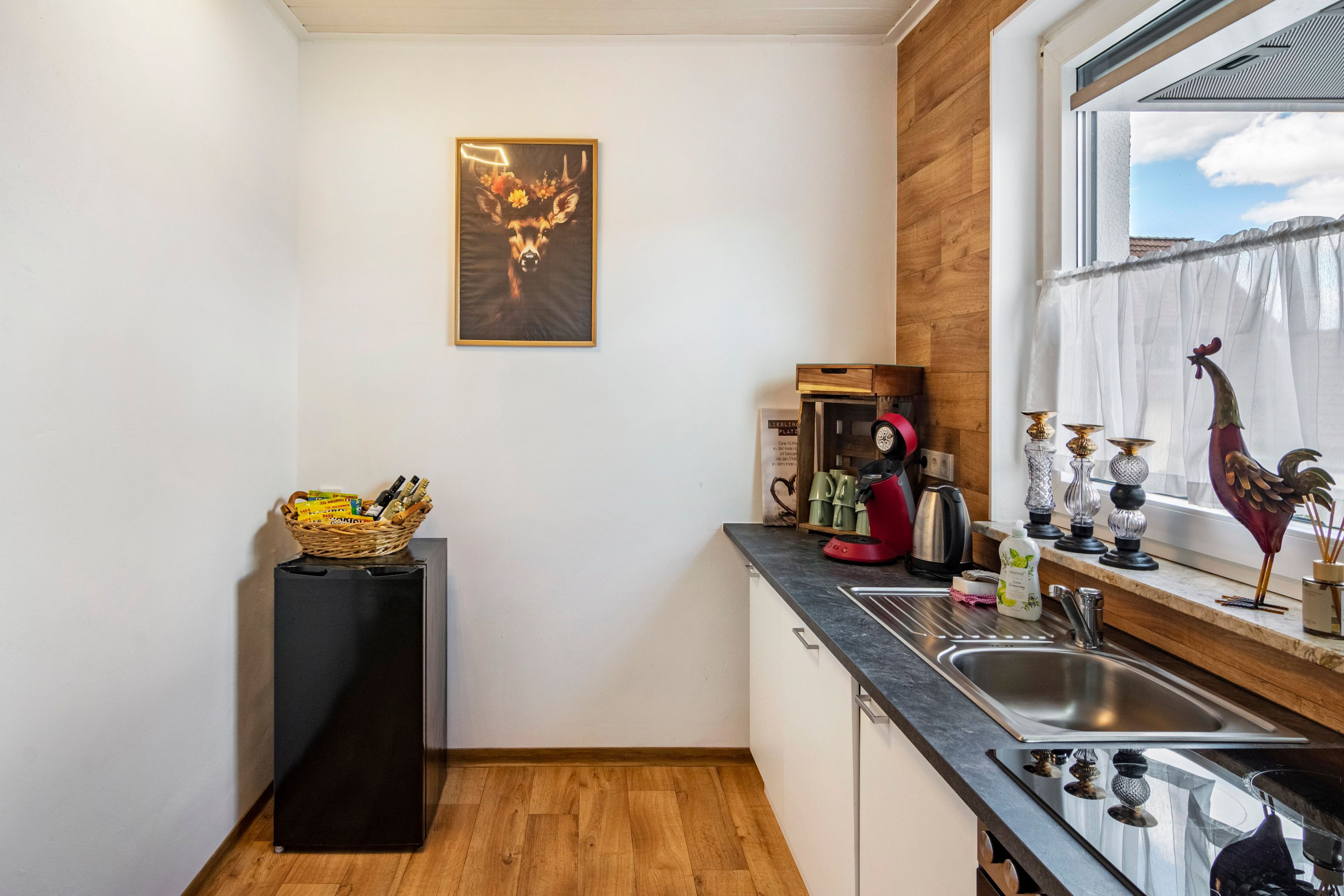 Une cuisine avec un plancher en bois, un réfrigérateur noir, un évier et une fenêtre donnant sur le ciel.