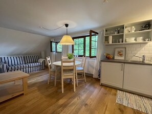 A cozy living room with a striped sofa, a dining table with chairs, and a kitchen area with shelves and a window.