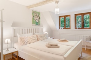 A bedroom with a bed, bedside tables, a lamp, a painting, and windows with a view of greenery.