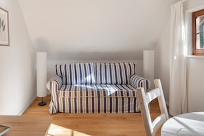 A room with a striped sofa, a white chair, a wooden floor, and a window with curtains.