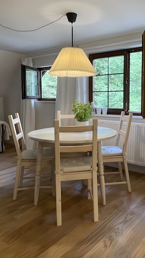 A dining area with a round table, four wooden chairs, a hanging lamp, and a window with a view of greenery.