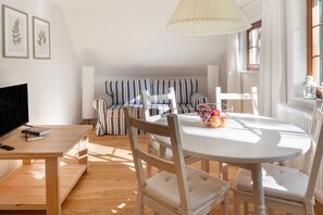 Een gezellige woonkamer met een gestreepte bank, een eettafel met stoelen, een tv op een dressoir en een raam met gordijnen.