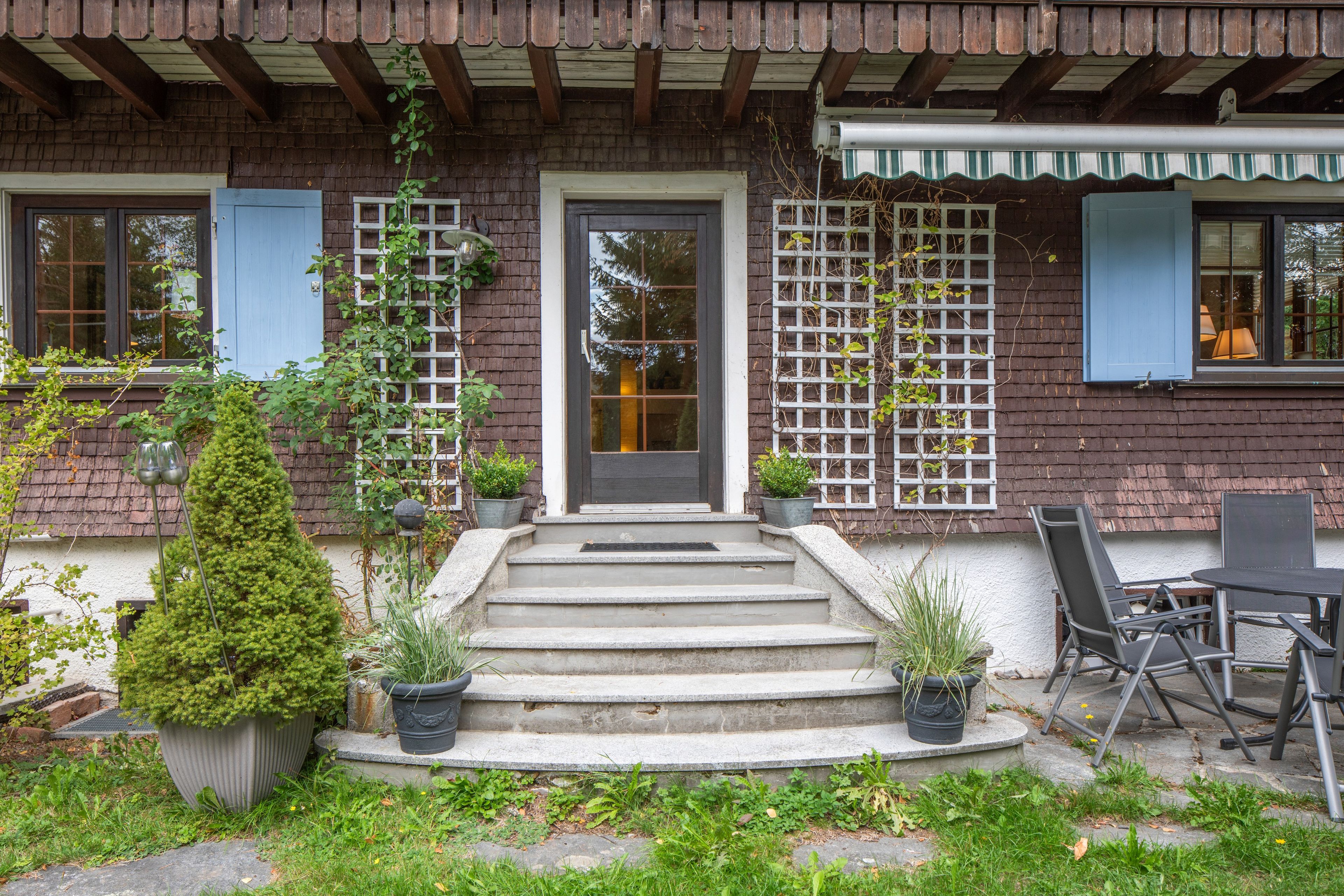 A house with a front door, steps leading up to it, a small garden with potted plants, and a patio area with outdoor furniture.