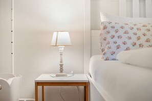 Una cama bien hecha con ropa de cama floral, una lámpara de noche sobre una mesita de madera y una pared blanca.