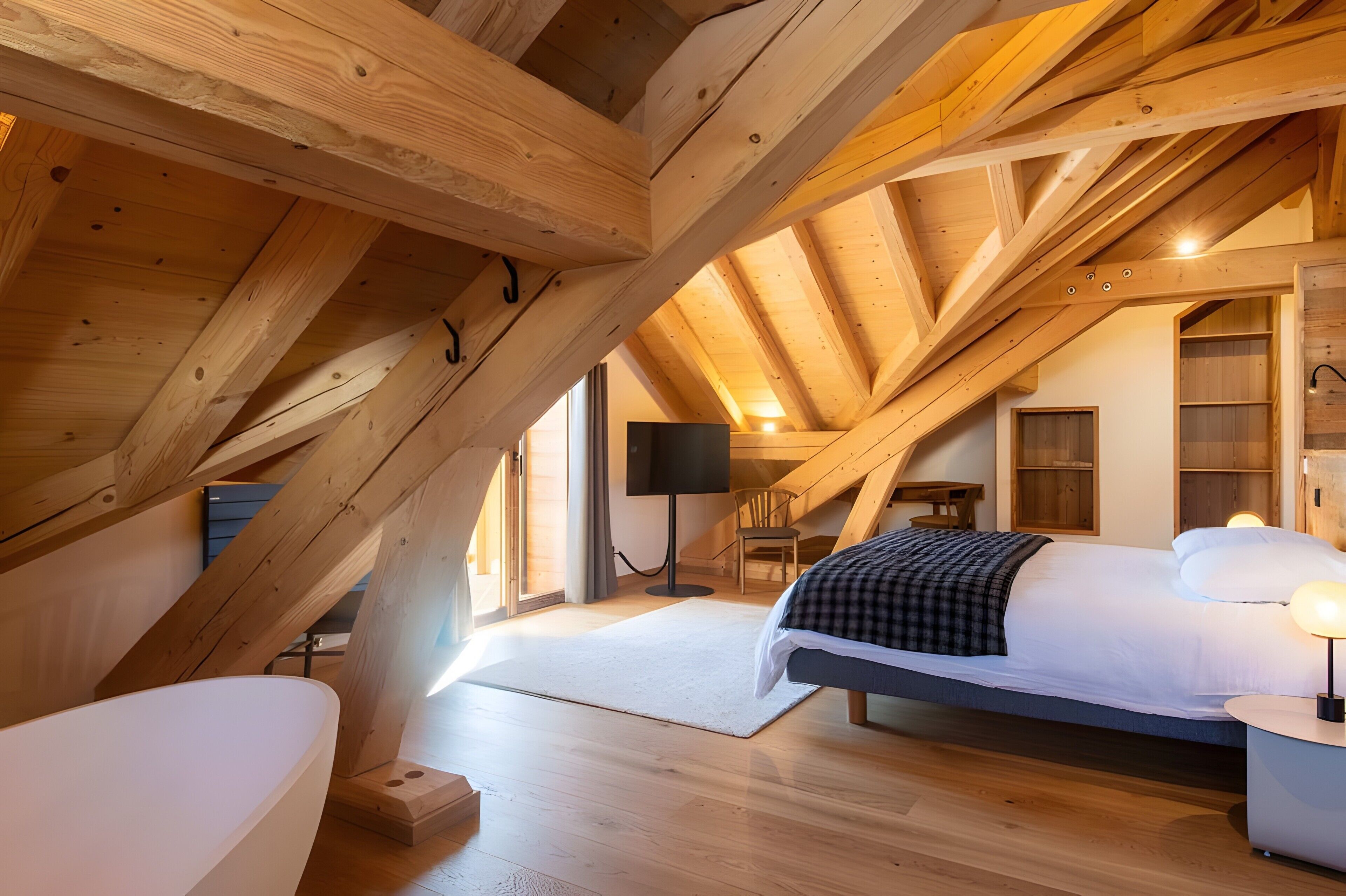 Une chambre avec un plafond en pente, des poutres en bois, un lit, une table de chevet, une lampe et un téléviseur.