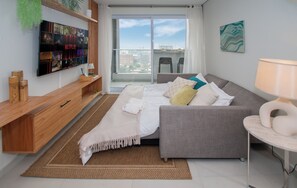 A modern living room with a grey sofa, a flat-screen TV mounted on the wall, a wooden side table, and a view of the city through large windows. - Corporate Stays First Living - 903 (Asunción)