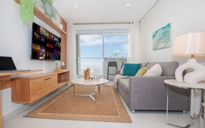A modern living room with a flat-screen TV, a grey sofa, a coffee table, and a view through a large window. - Corporate Stays First Living - 903 (Asunción)