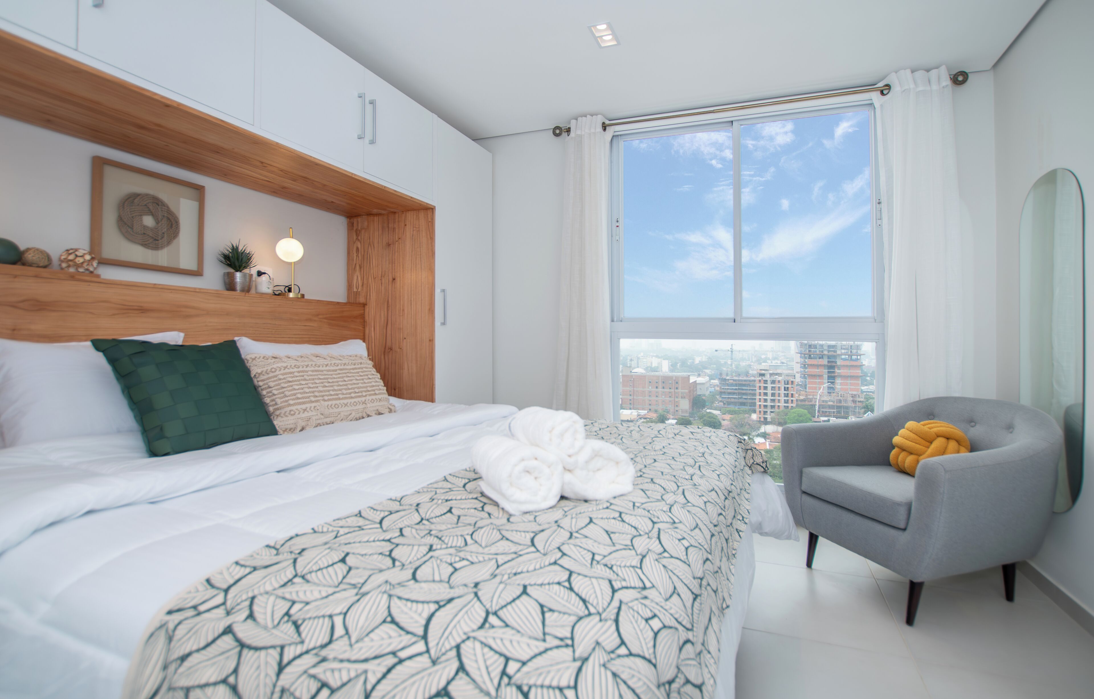 A modern hotel room with a large bed, a grey armchair, and a city view.