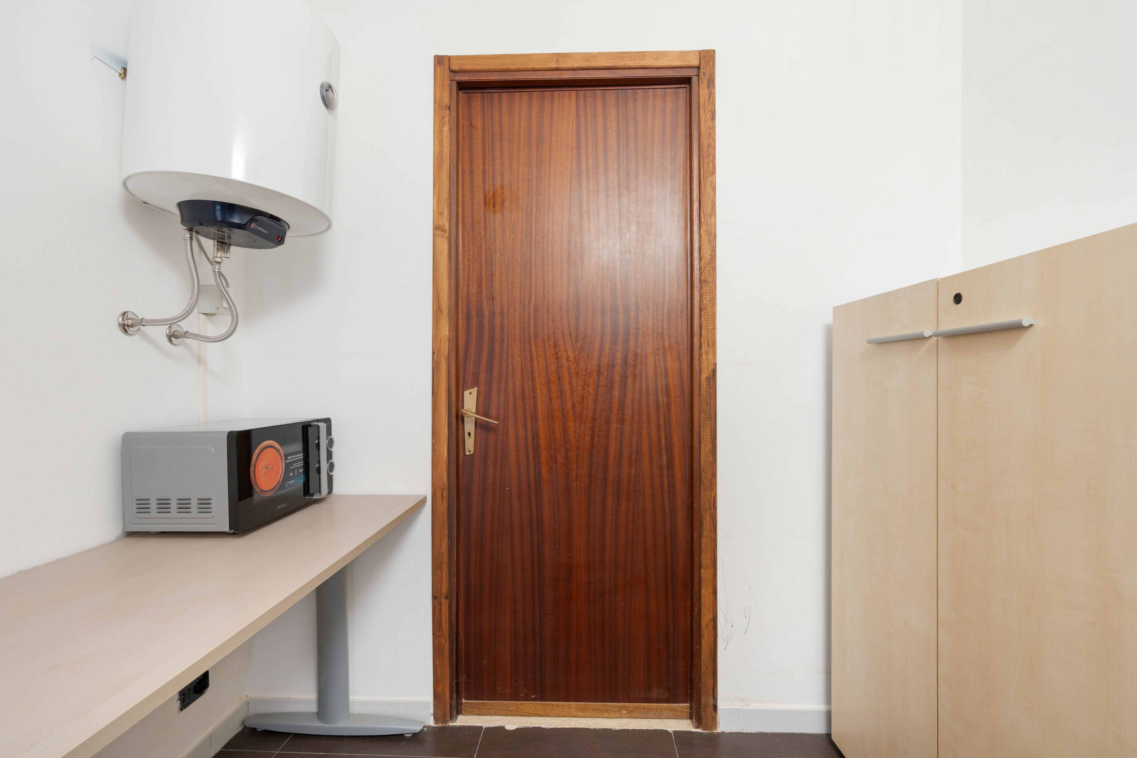 Een kamer met een houten deur, een witte muur, een grijze plank met een zwart apparaat, en een witte wandverwarmers.