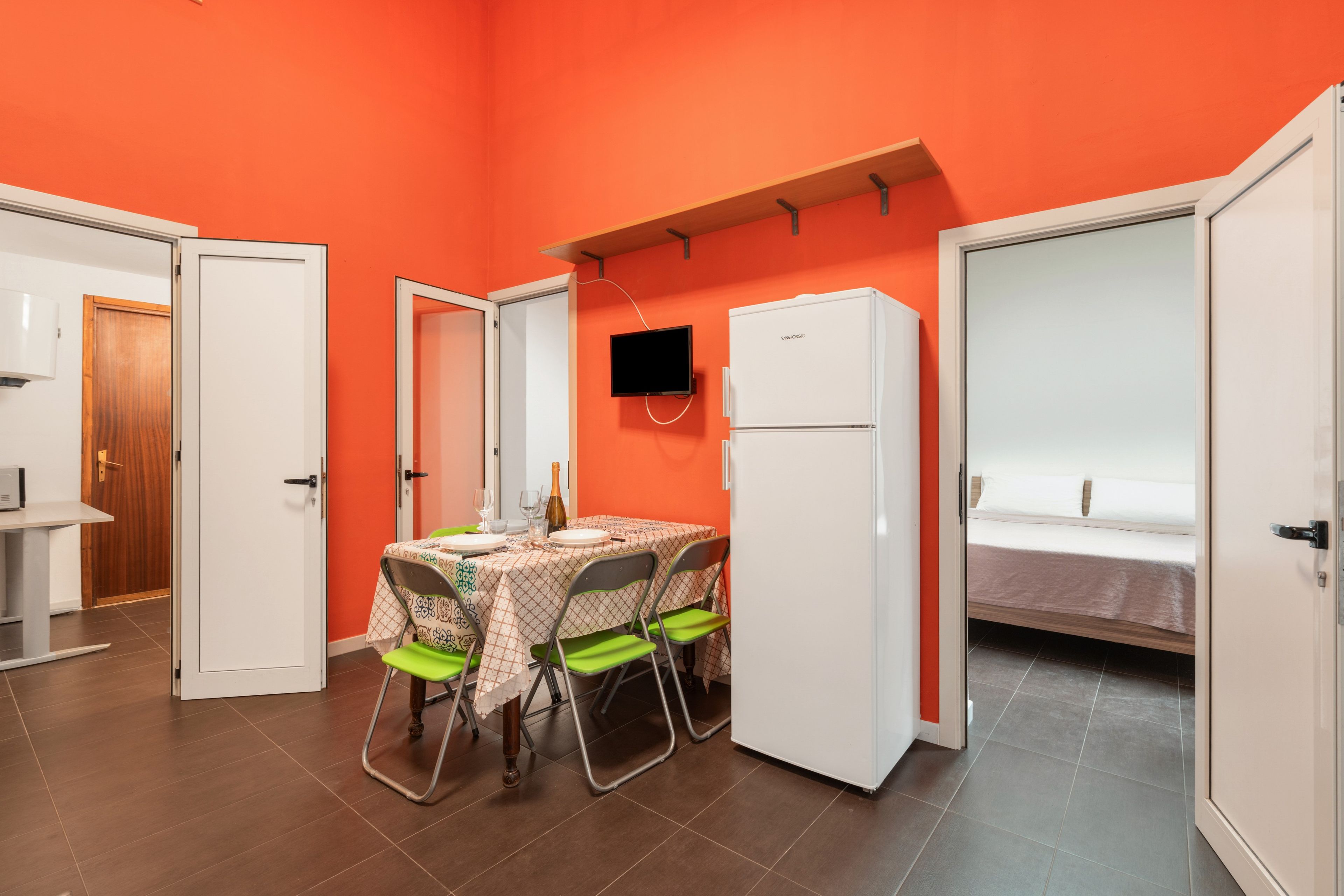 Une pièce de taille modeste comprenant une table à manger, des chaises, un réfrigérateur et un téléviseur fixé au mur.