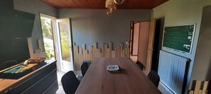 A room with a wooden table, chairs, a blackboard, and a window with a view of greenery. - Holiday Home 'La Chaumière Du Tank' with Private Terrace, Private Garden and Wi-Fi (Commes)