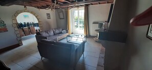 Ein Wohnzimmer mit einer Couch, einem Couchtisch und einem Fernseher. Es gibt ein Fenster mit Vorhängen, eine Heizung und eine Pendelleuchte.