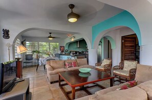 A spacious living area with a sofa, armchairs, a coffee table, and a television. There is a kitchen visible in the background with a dining area. - Wind Dancer - Ocean View Retreat (Vieques)