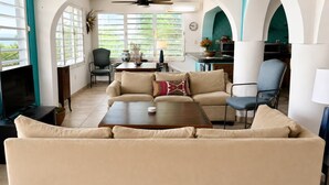 Een woonkamer met een bank, salontafel en een televisie. Er is een eethoek met een tafel en stoelen. De kamer heeft grote ramen en een plafondventilator.