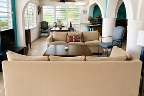A living room with a sofa, coffee table, and a television. There is a dining area with a table and chairs. The room has large windows and a ceiling fan. - Wind Dancer - Ocean View Retreat (Vieques)