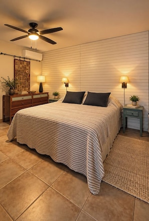 A bedroom with a bed, two bedside tables with lamps, a ceiling fan, a wall-mounted air conditioner, and a decorative mirror. - Guesthouse with Panoramic Ocean Views on a quiet street in Vieques (Vieques)