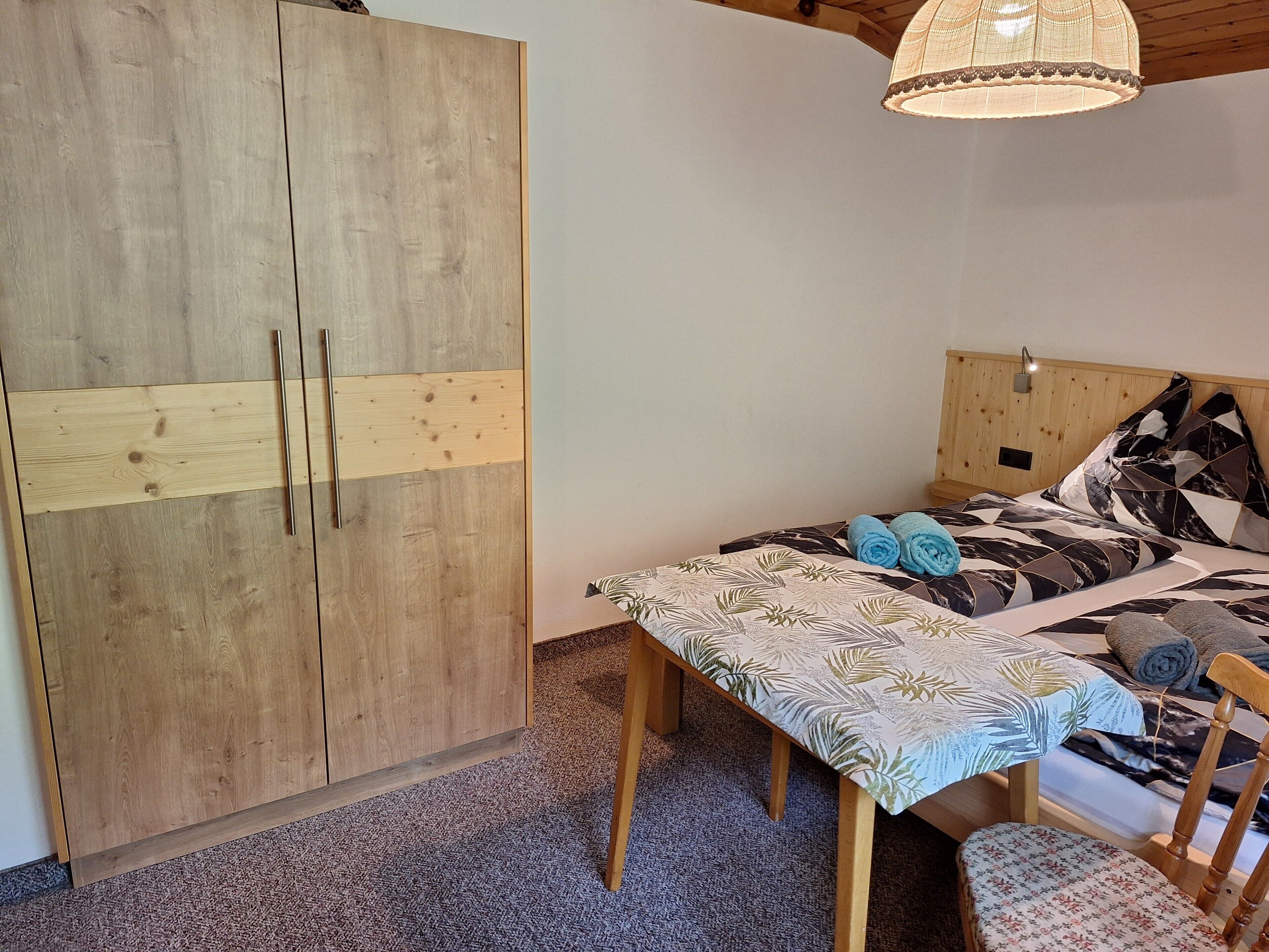 A bedroom with a wooden wardrobe, a bed with patterned bedding, a small table, and a chair.