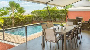 Ein Poolbereich im Freien mit einer überdachten Terrasse, Sitzgelegenheiten und Blick auf die umliegende Grünfläche.