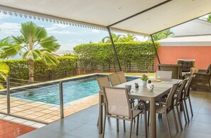 Ein Poolbereich im Freien mit einer überdachten Terrasse, Sitzgelegenheiten und Blick auf die umliegende Grünfläche.