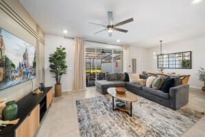 Un salon avec un canapé, une table basse et un téléviseur. On y trouve également un ventilateur de plafond, une plante et une fenêtre avec des rideaux.