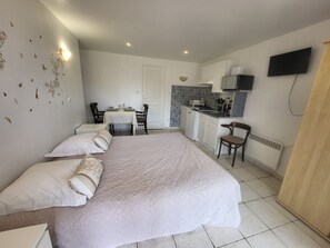 A bedroom with a bed, pillows, a bedside table, a small table with chairs, a kitchenette, and a TV mounted on the wall.