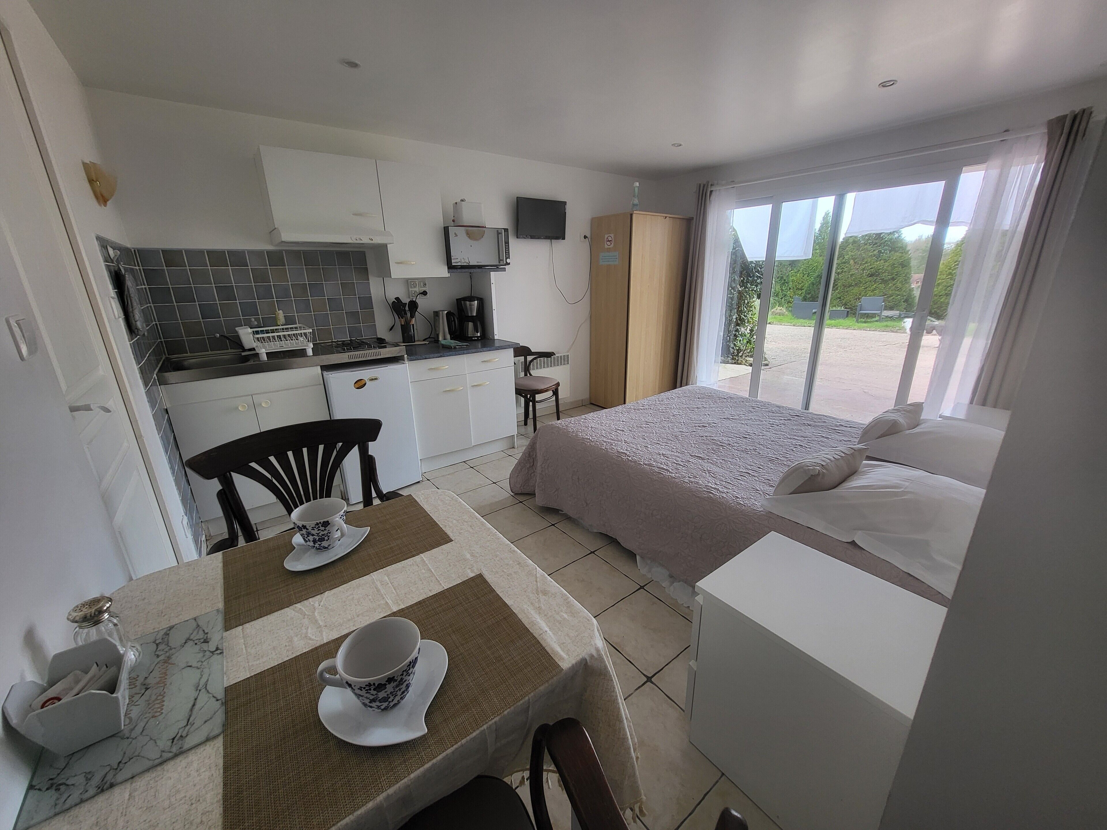 A compact hotel room with a kitchenette, a bed with a white comforter, and a large window offering a view of the outdoors.