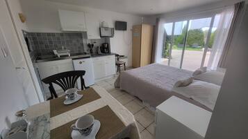 A compact hotel room with a kitchenette, a bed with a white comforter, and a large window offering a view of the outdoors.