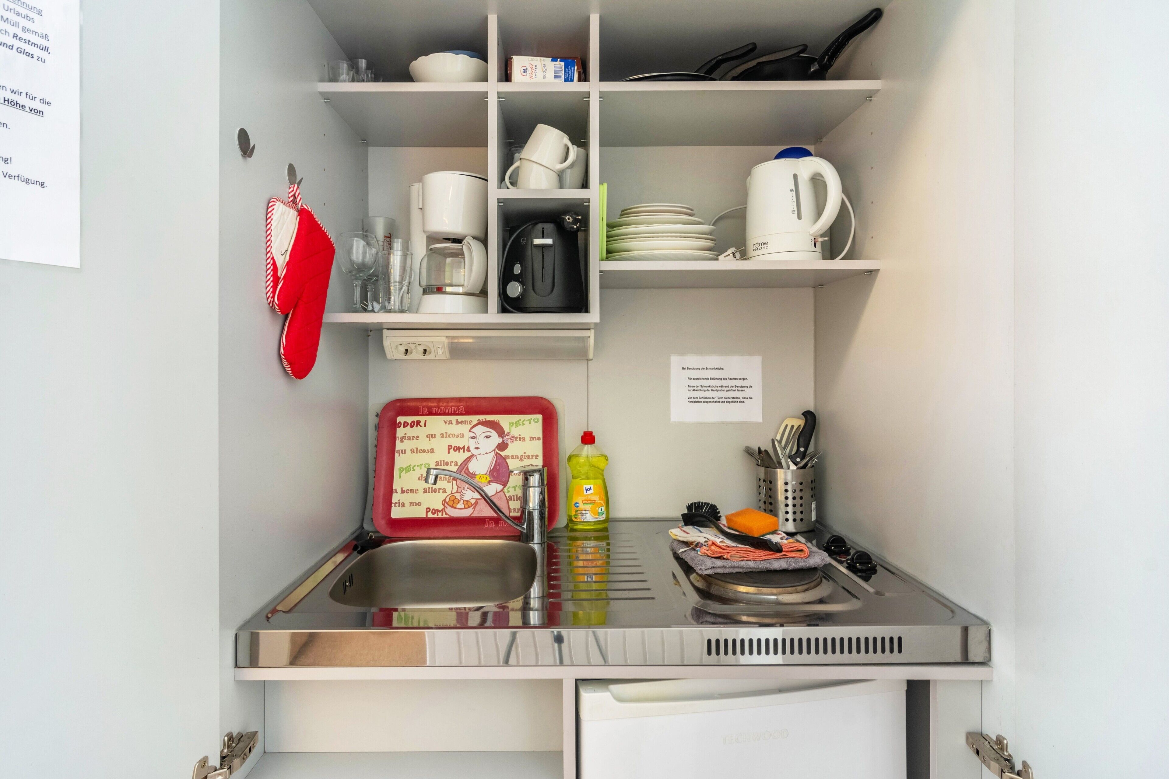Una kitchenette compacta con un fregadero, cafetera, hervidor y diversos utensilios de cocina.