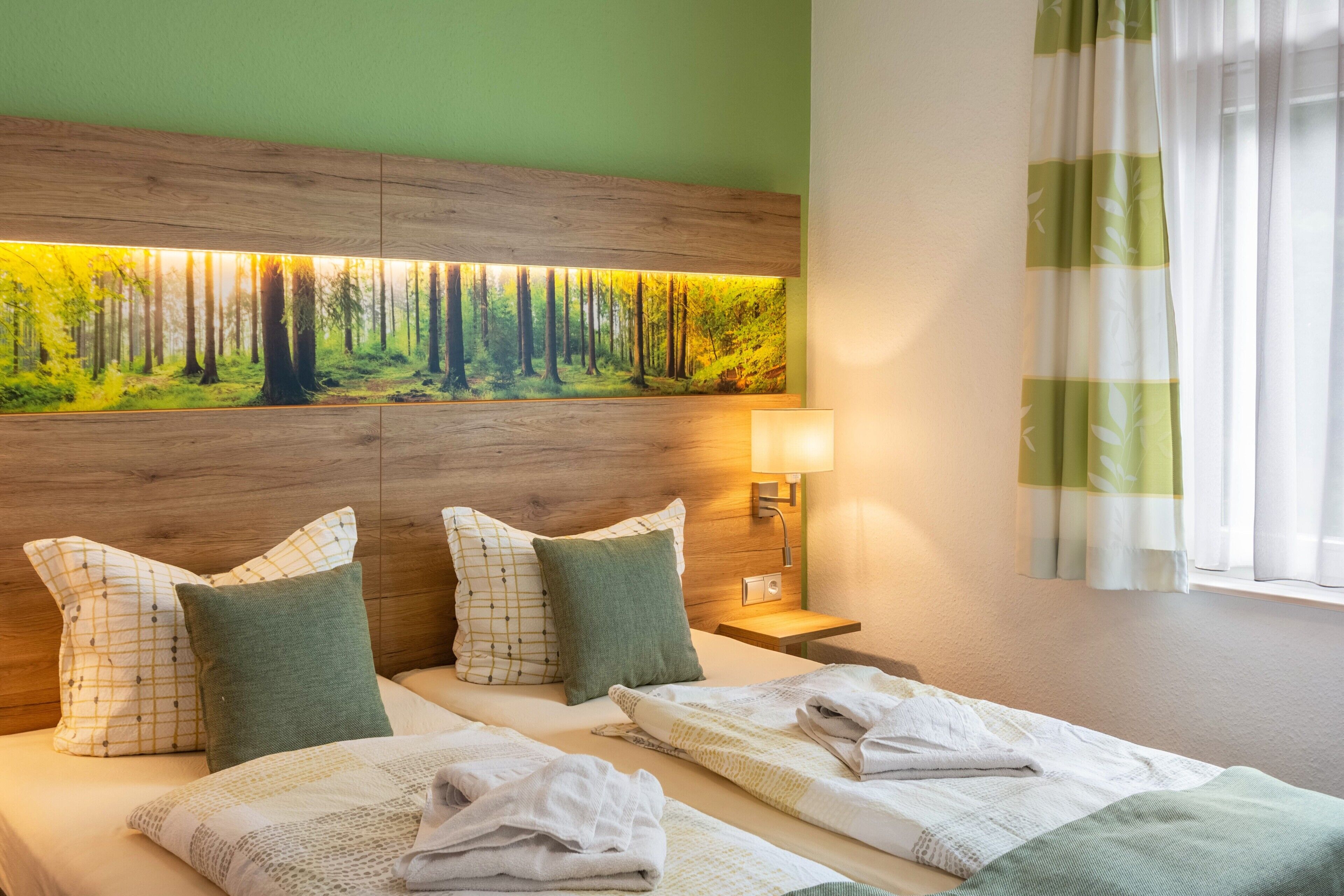 A hotel room with two beds, a large forest-themed wall art, a bedside lamp, and a window with sheer curtains.