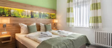 A hotel room with a bed, two bedside tables, a nightstand, a window with curtains, and a wall with a nature-themed mural.