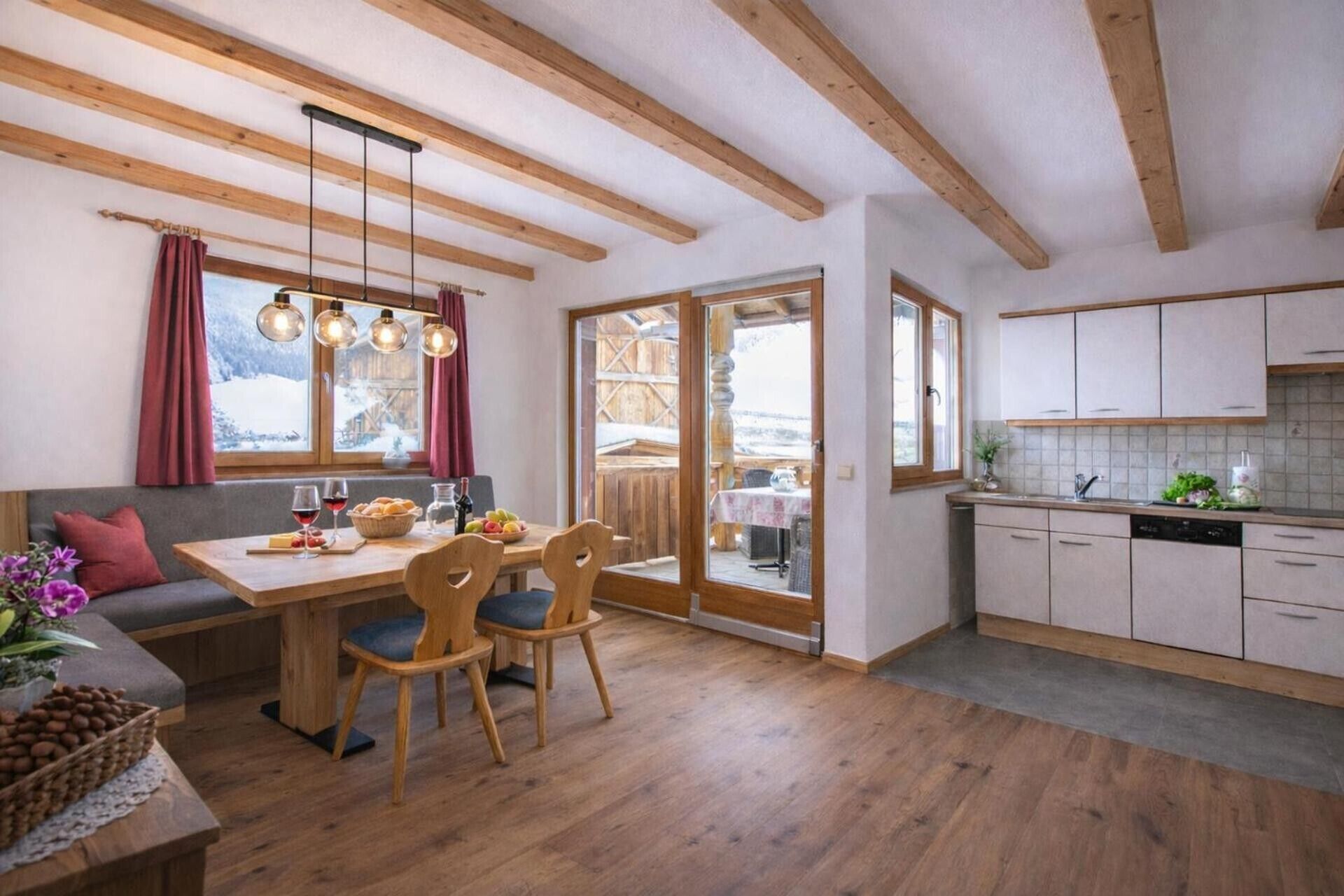A cozy kitchen with wooden beams, a dining area with a table and chairs, and a view of a snowy landscape through the windows.