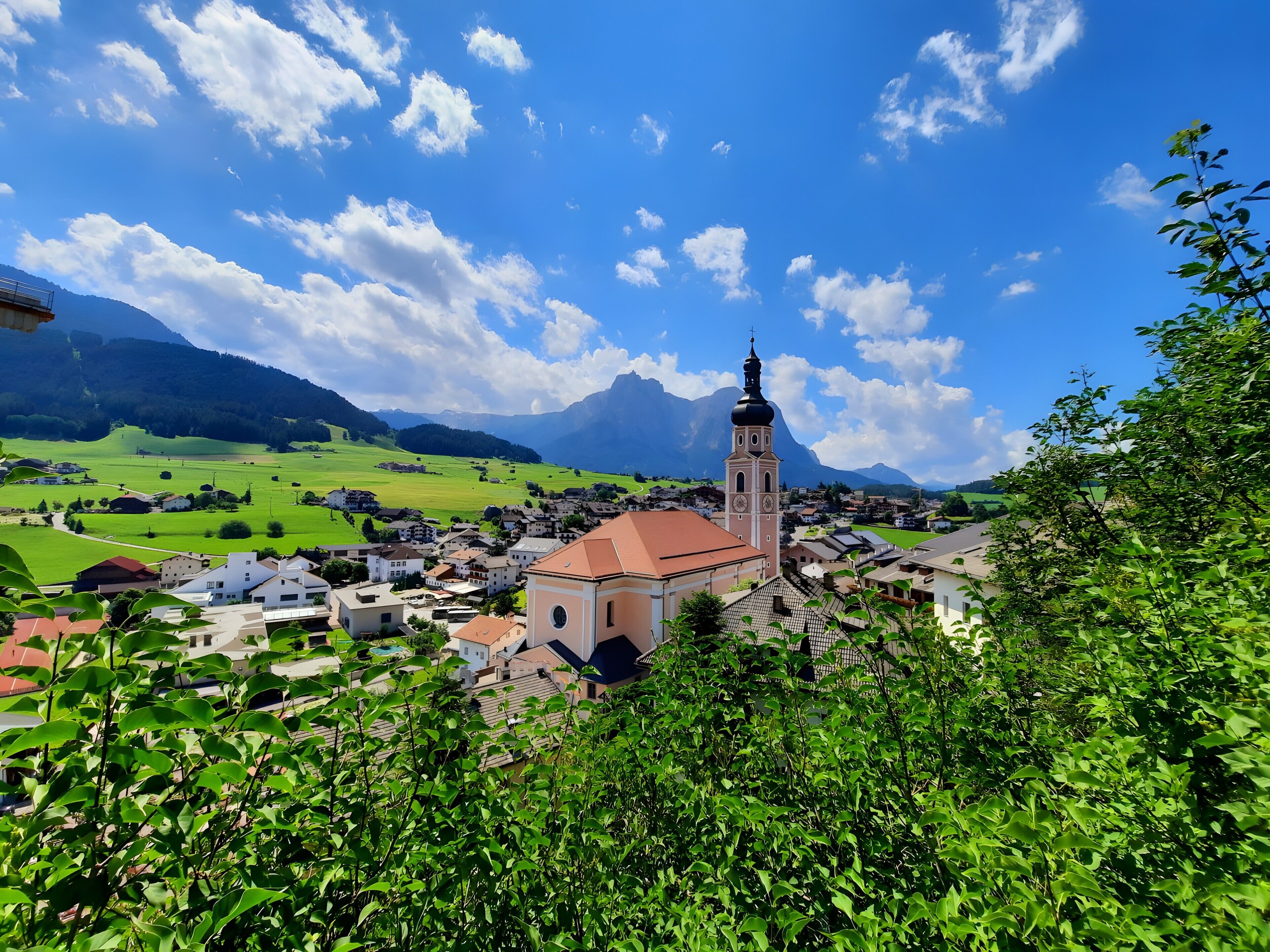 A town with a prominent church tower, surrounded by green fields and mountains.