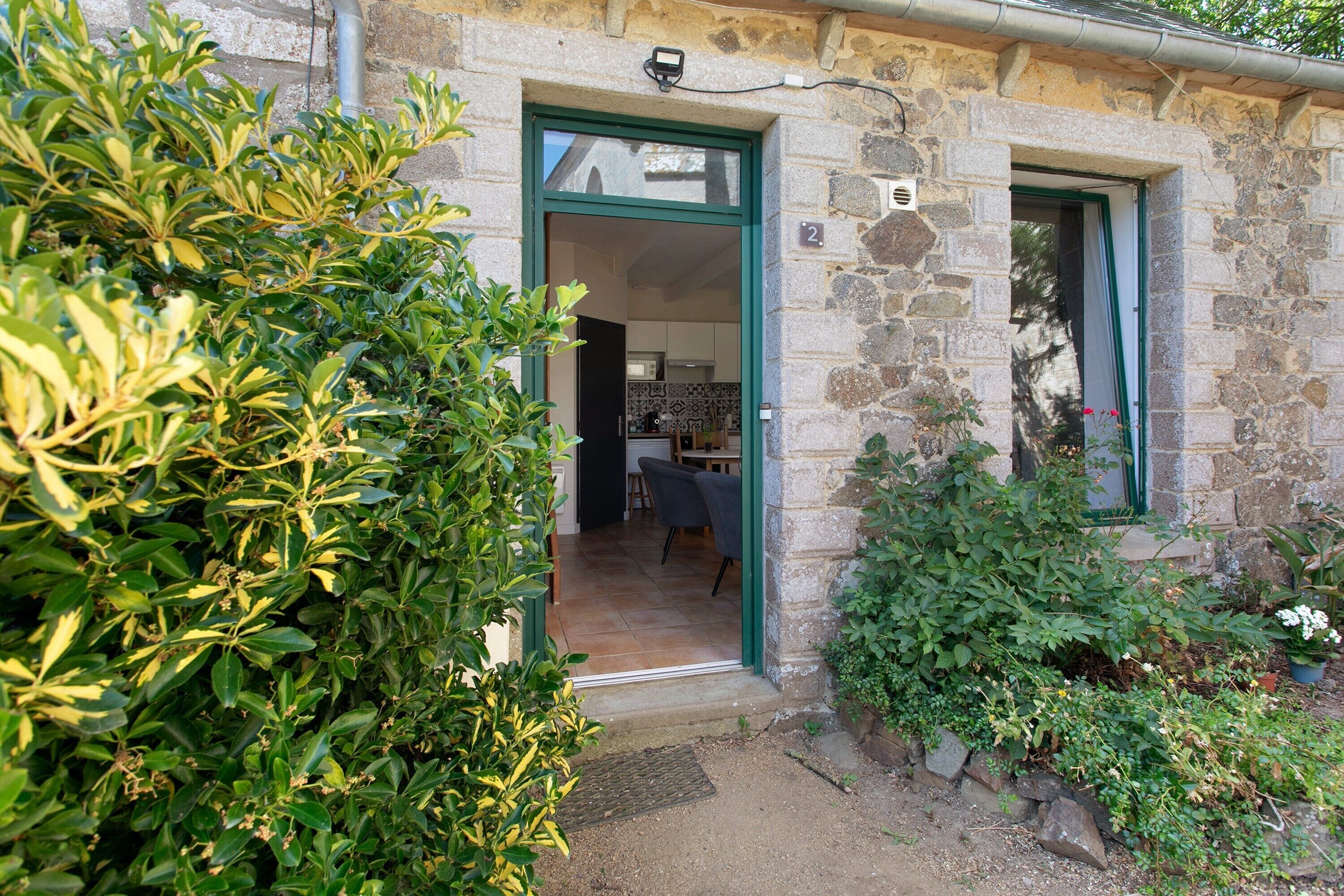 A stone building with a green door, a visible kitchen area, and a garden with various plants.