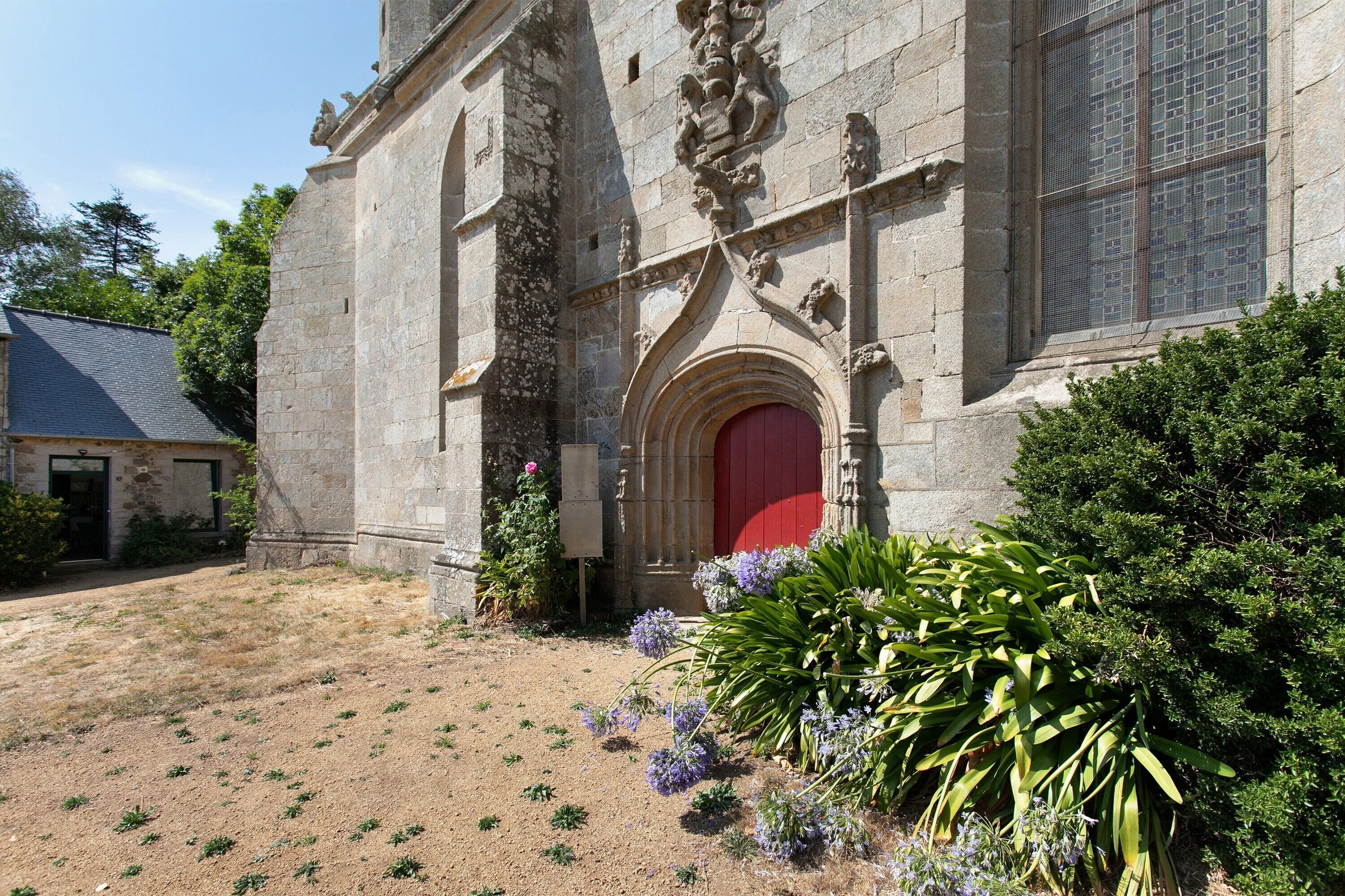 A stone building with a red door, surrounded by greenery and a clear sky.
