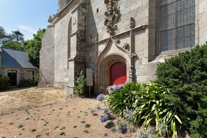 A stone building with a red door, surrounded by greenery and a clear sky.