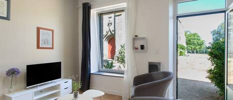 A modern living room with a flat-screen TV, a small white table, a grey armchair, and a window with a view of a courtyard.