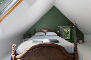 A bedroom with a sloped ceiling, a wooden bed, a nightstand, a lamp, and a window with curtains.