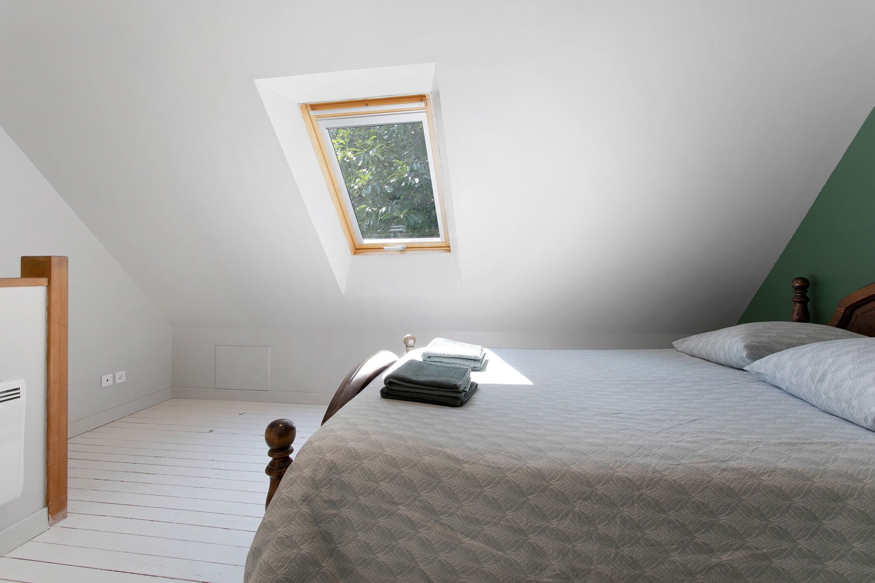 Un dormitorio con techo inclinado, una cama con colcha gris estampada, cabecera de madera y una ventana pequeña con vistas a la vegetación.