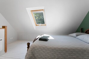 A bedroom with a sloped ceiling, a bed with a grey patterned bedspread, a wooden headboard, and a small window with a view of greenery.