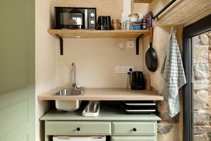 Una cocina compacta con microondas, hervidor de agua y cafetera sobre una estantería, un lavabo con grifo y una pared de piedra.