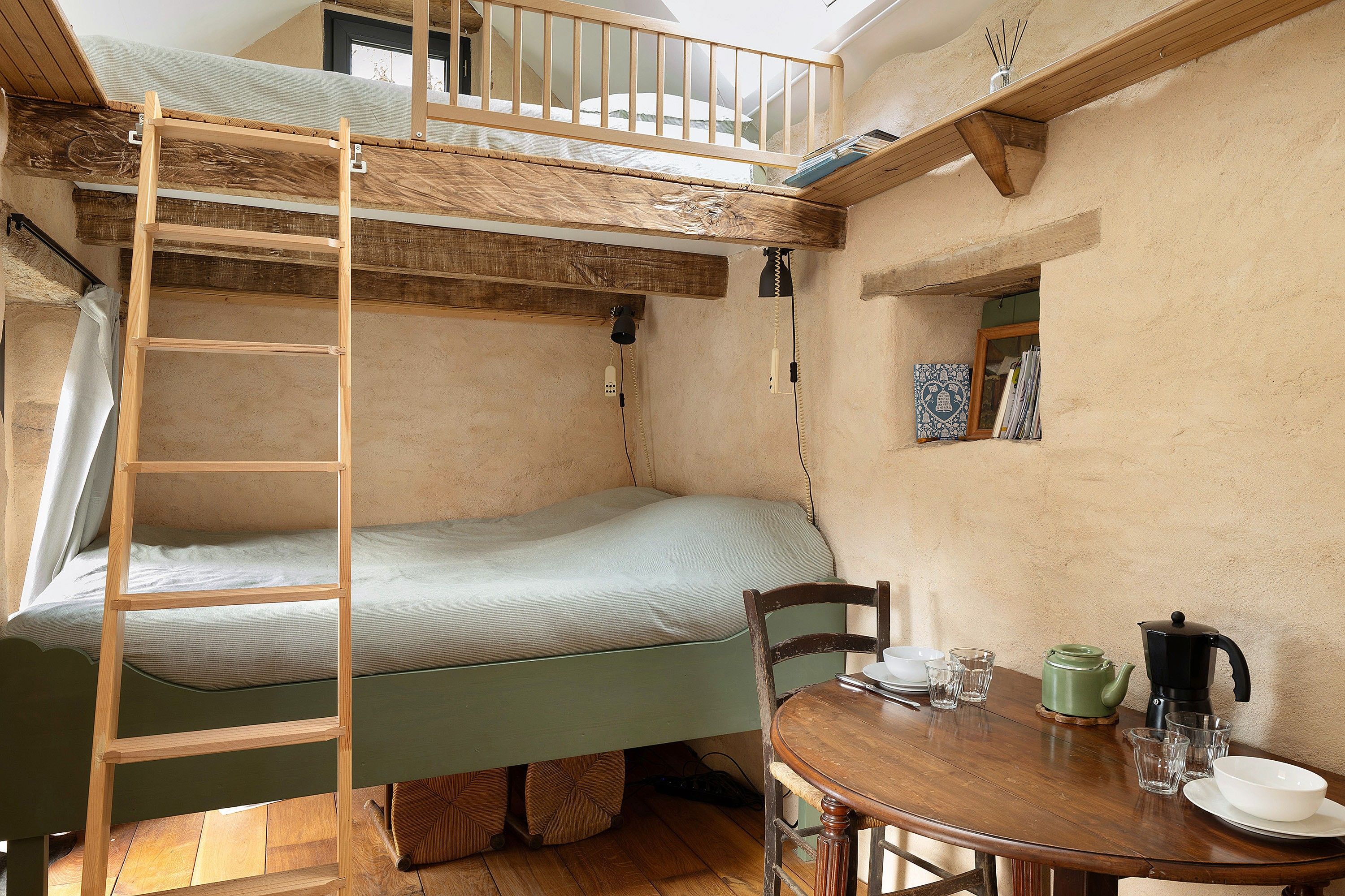 A bunk bed with a ladder, a table with a kettle, cups, and a bowl, and a small window with a picture frame.