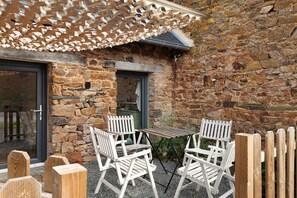 Un bâtiment en pierre avec une porte et une fenêtre en bois, des sièges extérieurs avec des chaises blanches et un auvent en treillis.