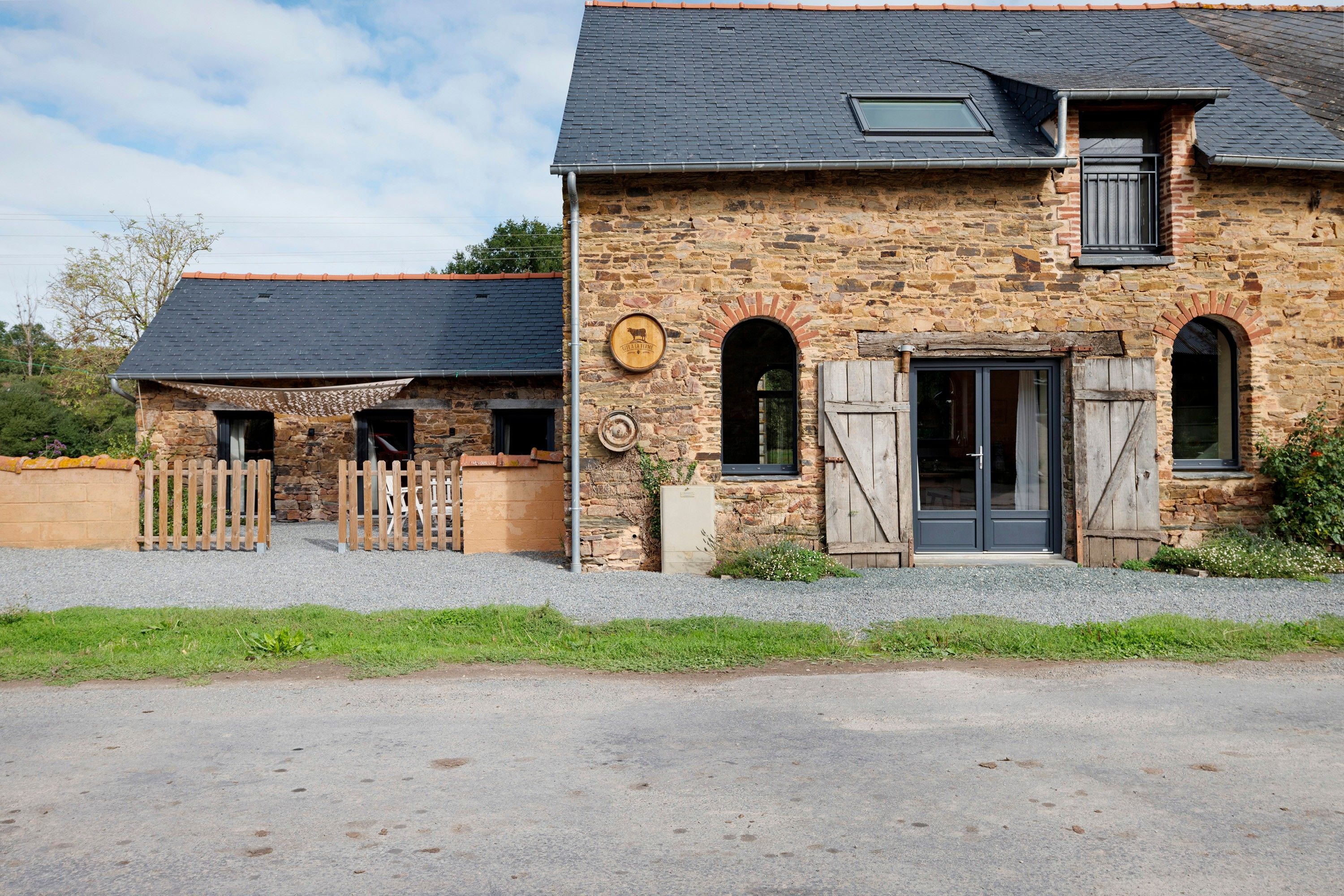 A stone building with a dark roof, a wooden fence, and a gravel driveway.
