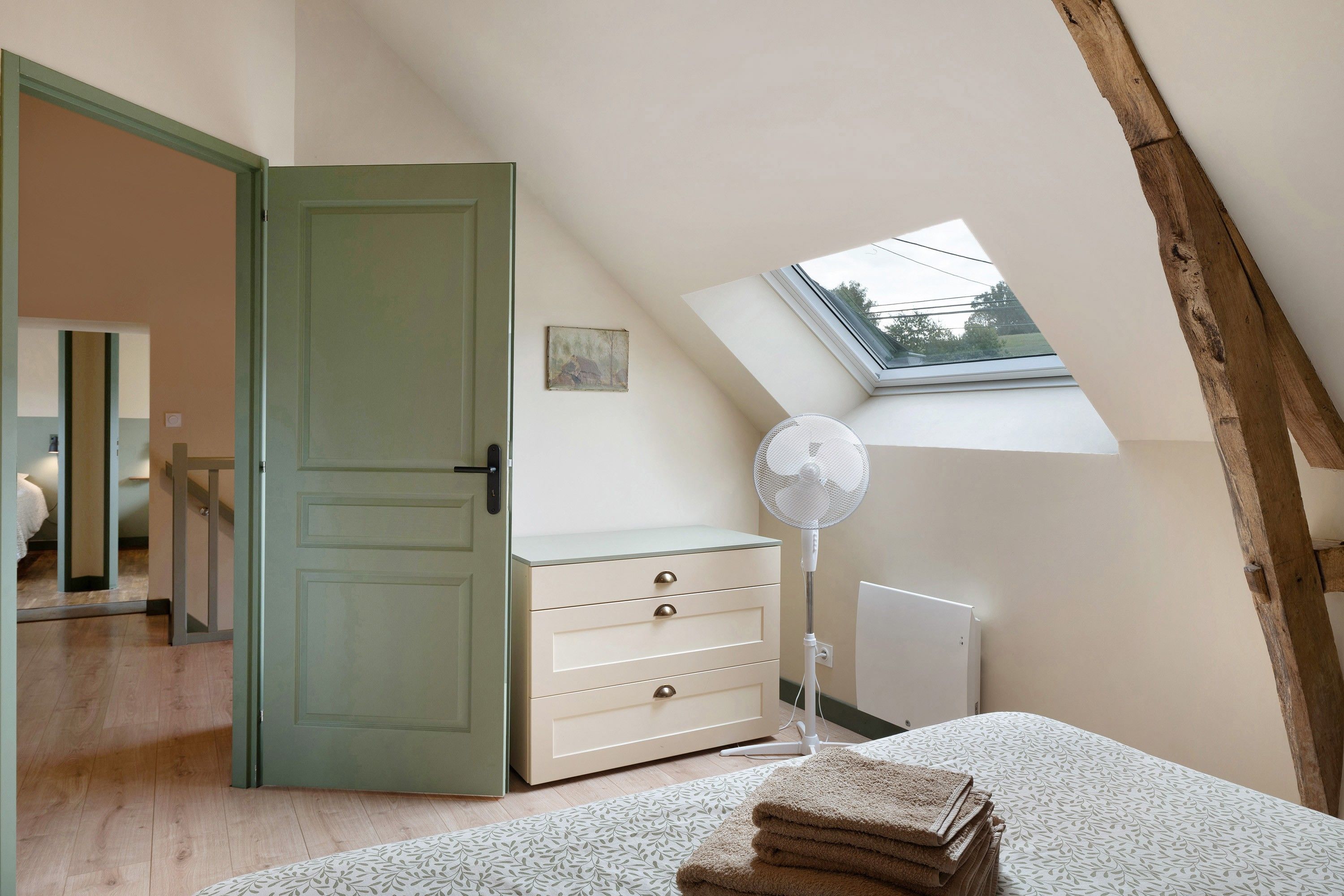 A bedroom with a sloped ceiling, a skylight, a bed with a patterned bedspread, a bedside table with a fan, and a dresser with drawers.