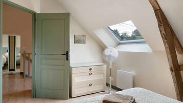 A bedroom with a sloped ceiling, a skylight, a bed with a patterned bedspread, a bedside table with a fan, and a dresser with drawers.