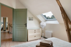 Ein Schlafzimmer mit schrägem Dach, einem Oberlicht, einem Bett mit gemustertem Überwurf, einem Nachttisch mit Ventilator und einer Kommode mit Schubladen.
